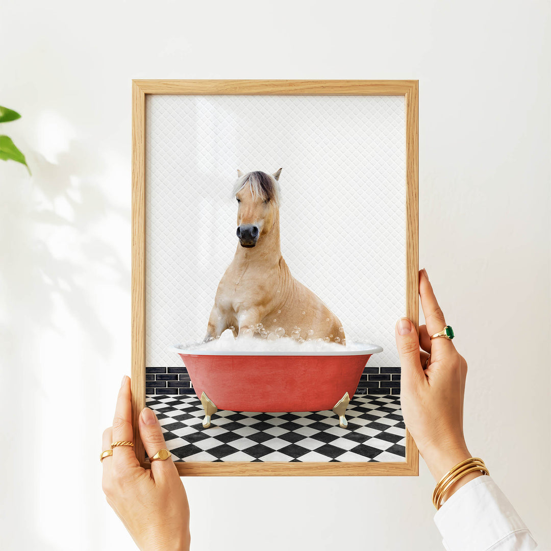 A person's hands are holding a framed picture of a horse sitting in a red bathtub filled with bubbles, with a black and white checkered floor in the background.