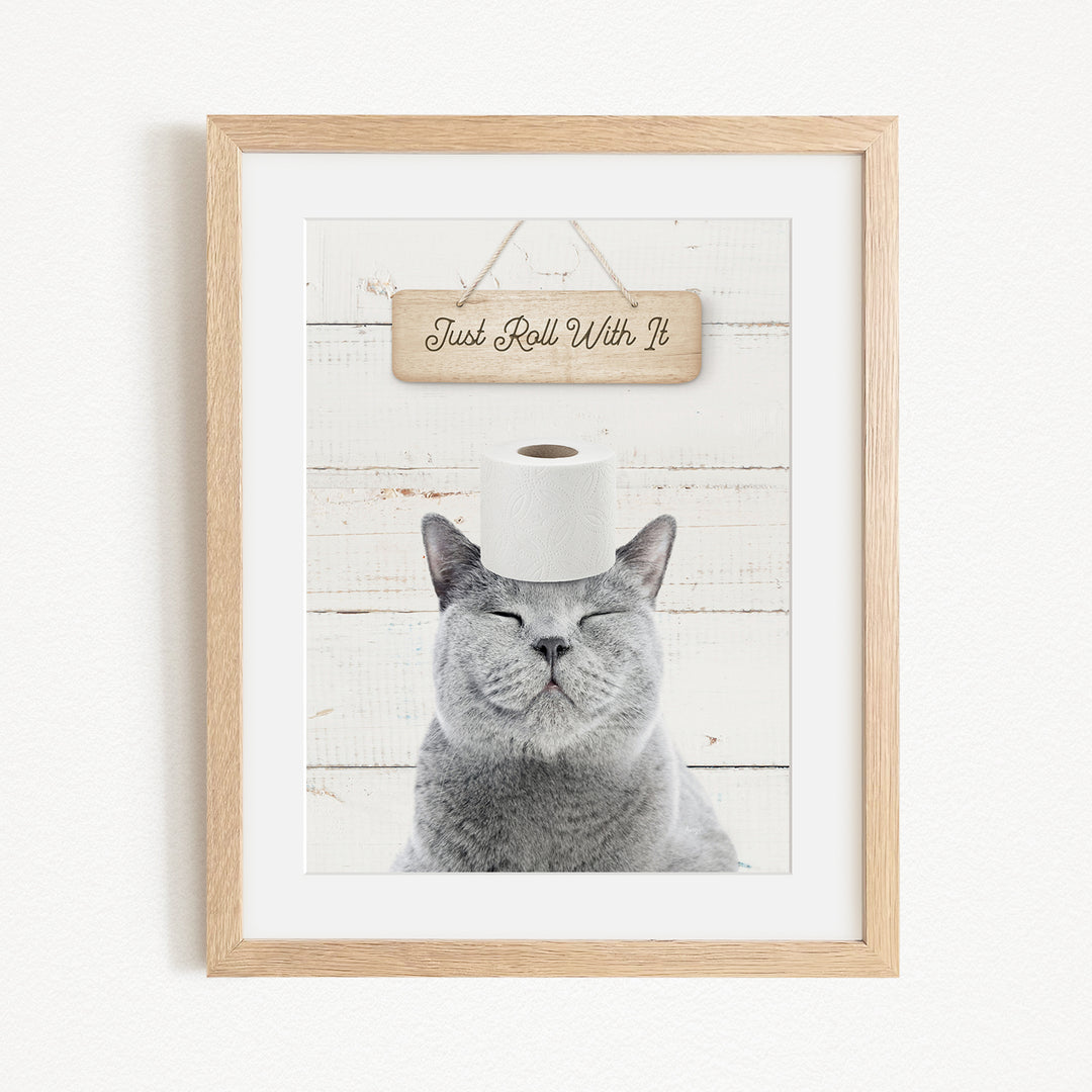 A framed image of a gray cat with its eyes closed, wearing a roll of toilet paper on its head, with a wooden sign above that says "Just Roll With It".
