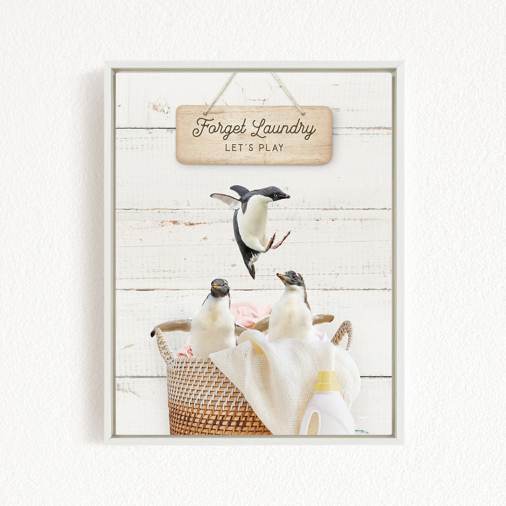 A framed image of three penguins in a basket, with a wooden sign above them that says "Forget Laundry, Let's Play".