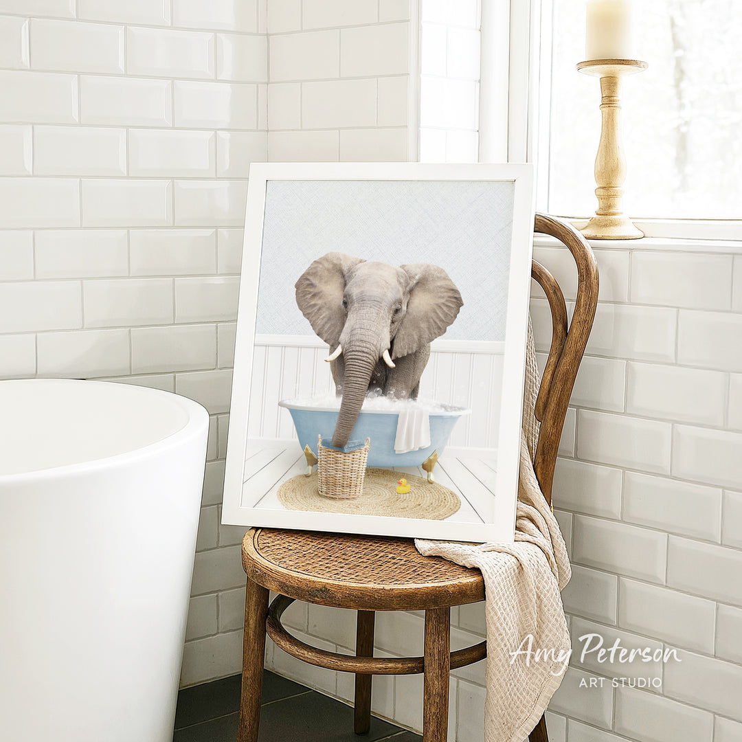 A framed image of an elephant sitting in a bathtub, surrounded by a basket of toys, is displayed on a wooden chair in a bathroom setting.