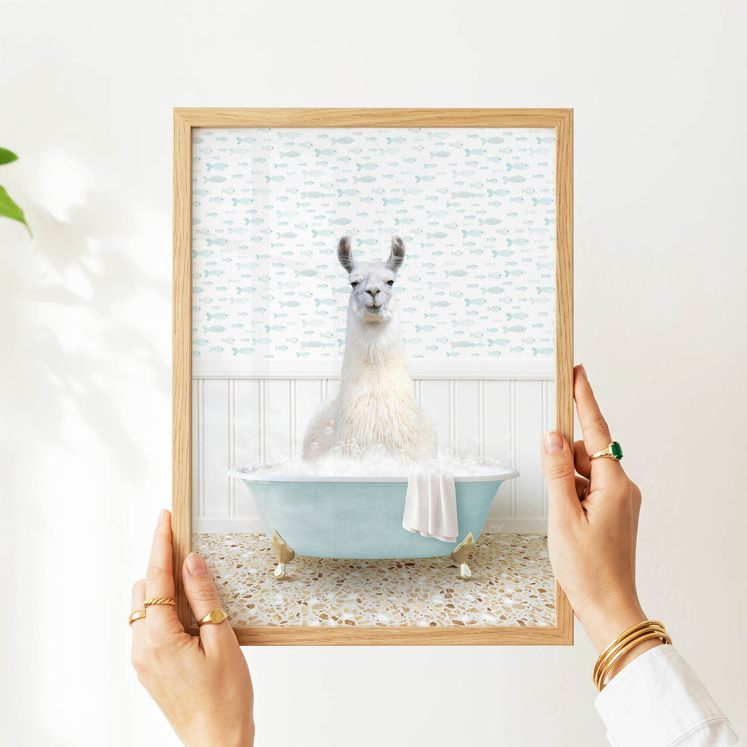 A person's hands are holding a framed picture of a llama sitting in a bathtub filled with bubbles, with a patterned wall behind it.