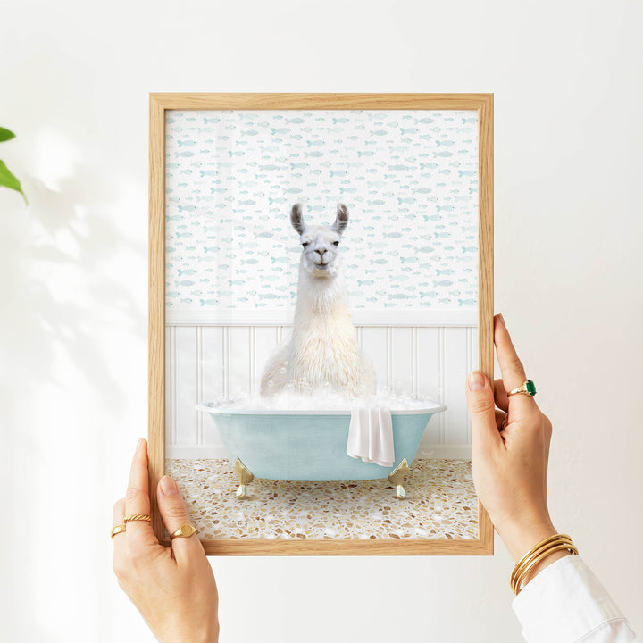 A person's hands are holding a framed picture of a llama sitting in a bathtub filled with bubbles, with a patterned wall behind it.