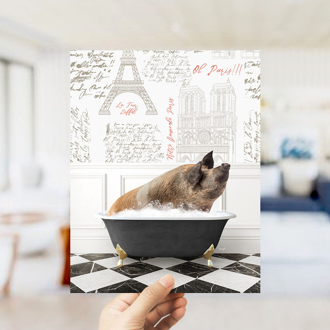 A hand holds up a printed image featuring a pig sitting in a bathtub filled with bubbles, with a Paris-themed background.