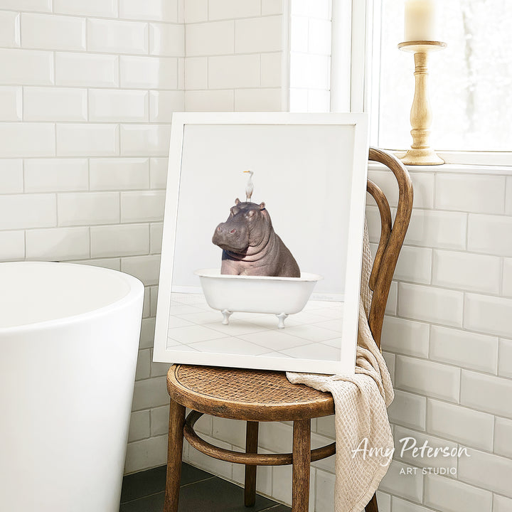 A framed image of a hippopotamus sitting in a bathtub is displayed on a wooden chair in a bathroom setting.