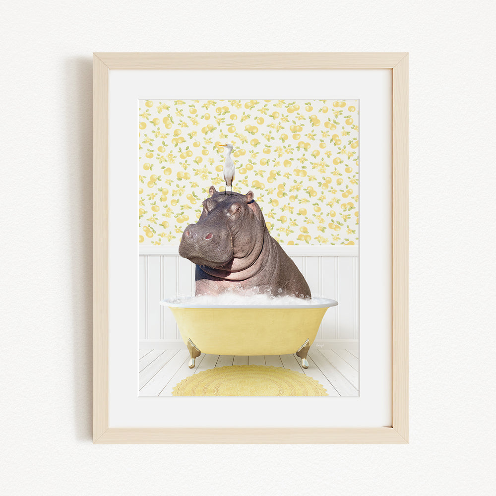 A framed image depicting a hippopotamus sitting in a yellow bathtub filled with water and bubbles, with a yellow and white floral patterned wall behind it.