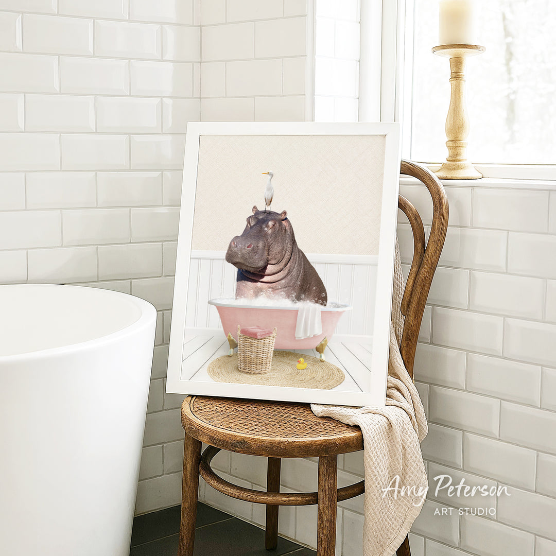 A framed image of a hippopotamus sitting in a pink bathtub, surrounded by a basket of toys and a towel, is displayed on a wooden chair in a bathroom setting.