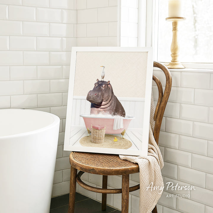 A framed image of a hippopotamus sitting in a pink bathtub, surrounded by a basket of toys and a towel, is displayed on a wooden chair in a bathroom setting.