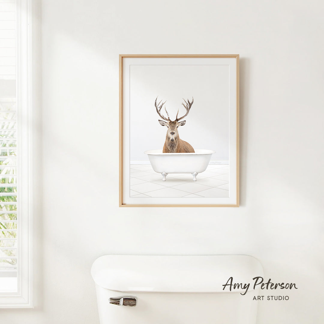 A framed image of a deer sitting in a bathtub is hanging on a white wall.