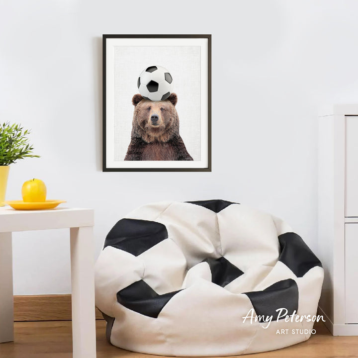 a framed picture of a bear balancing a soccer ball on its head, with a cozy bean bag chair in the foreground and a potted plant nearby.