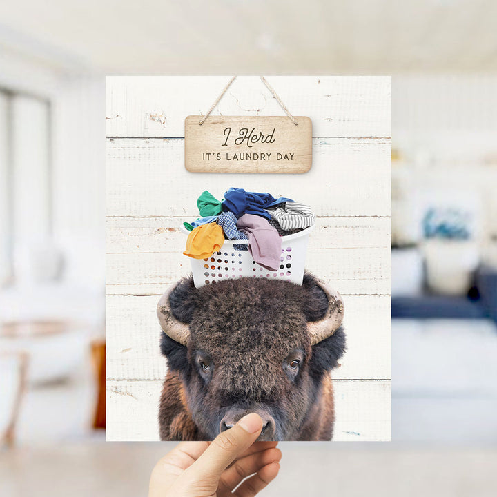 A hand holding a sign that says "I herd, it's laundry day" above a picture of a bison with clothes on its head.