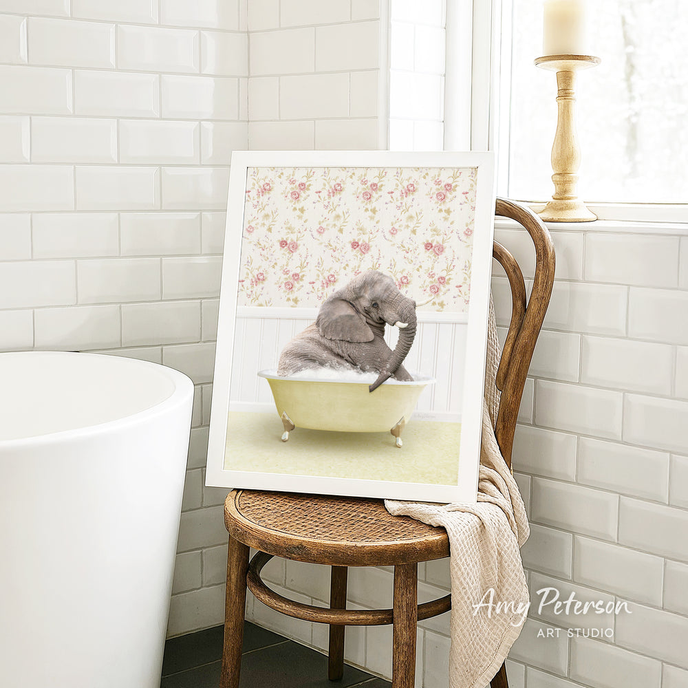 A framed image of a gray elephant sitting in a yellow bathtub, surrounded by a floral pattern, is displayed on a wooden chair in a bathroom setting.