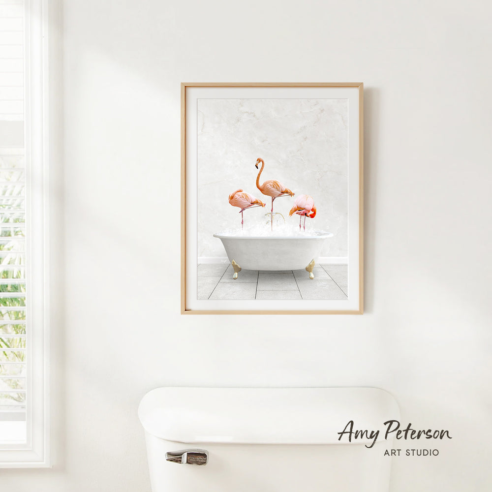 A framed print of three flamingos standing in a white bathtub is hanging on a white wall.