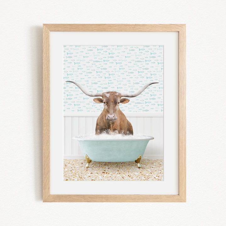 A framed image depicting a brown cow with large horns sitting in a light blue bathtub filled with bubbles.