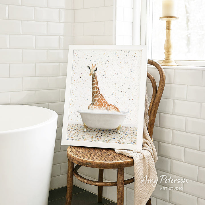A framed painting of a giraffe sitting in a bathtub is displayed on a wooden chair in a bathroom setting.