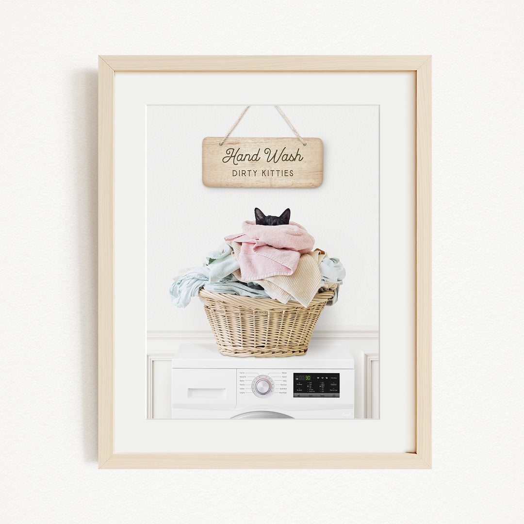 A framed image of a cat sitting in a basket filled with laundry, with a sign above that says "Hand Wash Dirty Kittens".