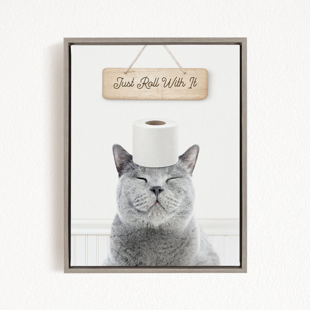 A gray cat with its eyes closed and a roll of toilet paper on its head, with a wooden sign above that says "Just Roll With It".