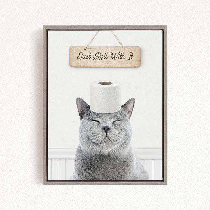 A gray cat with its eyes closed and a roll of toilet paper on its head, with a wooden sign above that says "Just Roll With It".