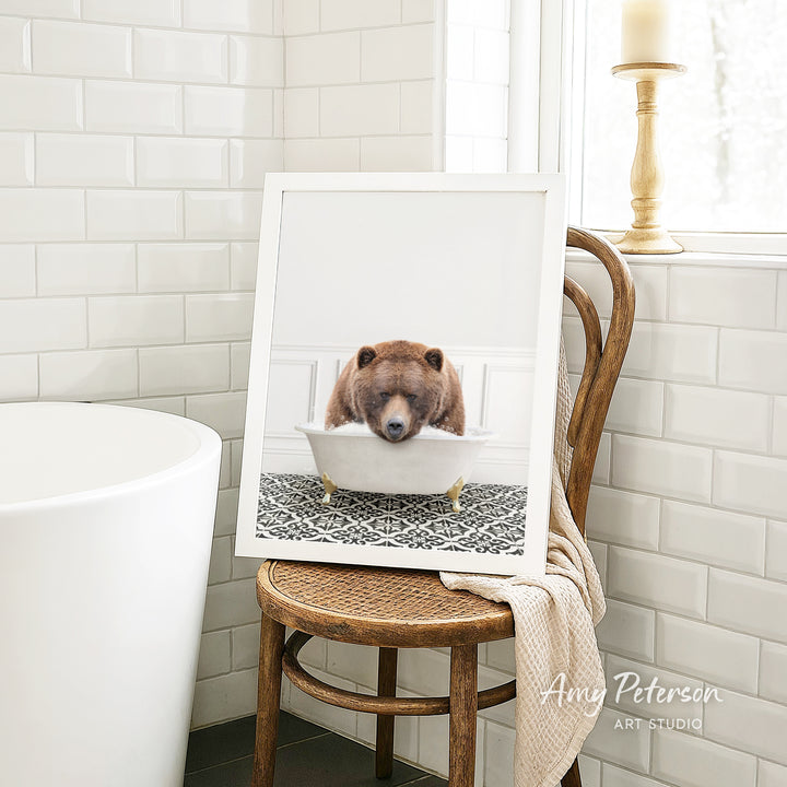 A framed image of a brown bear sitting in a bathtub, with a towel draped over the back of a wooden chair in a bathroom setting.