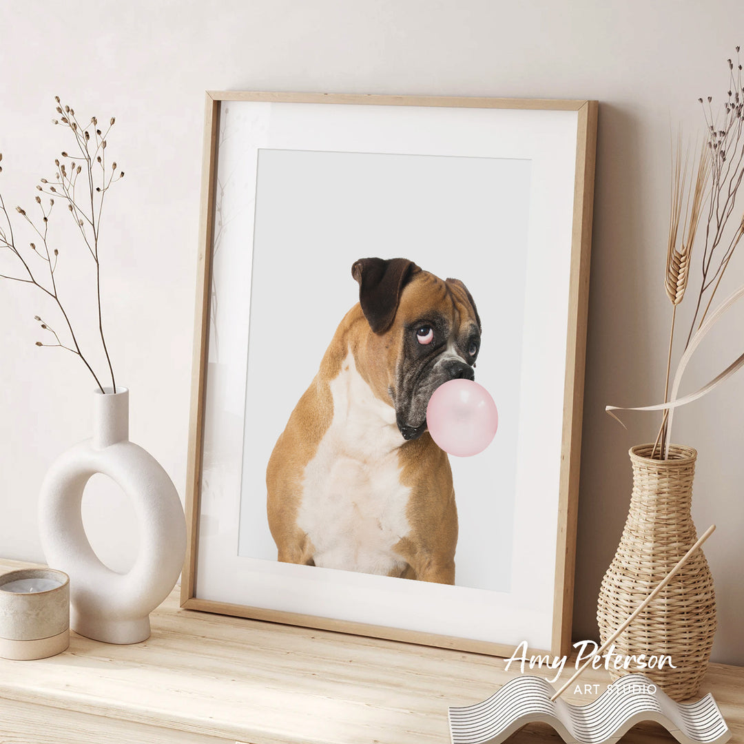 A framed photograph of a brown and white dog blowing a bubble with a pink bubble gum bubble, accompanied by decorative vases and a woven basket on a wooden surface.