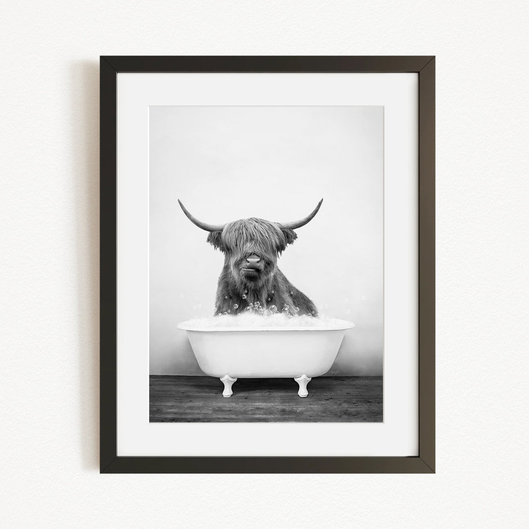 A black and white photograph of a Highland cow sitting in a bathtub filled with water.