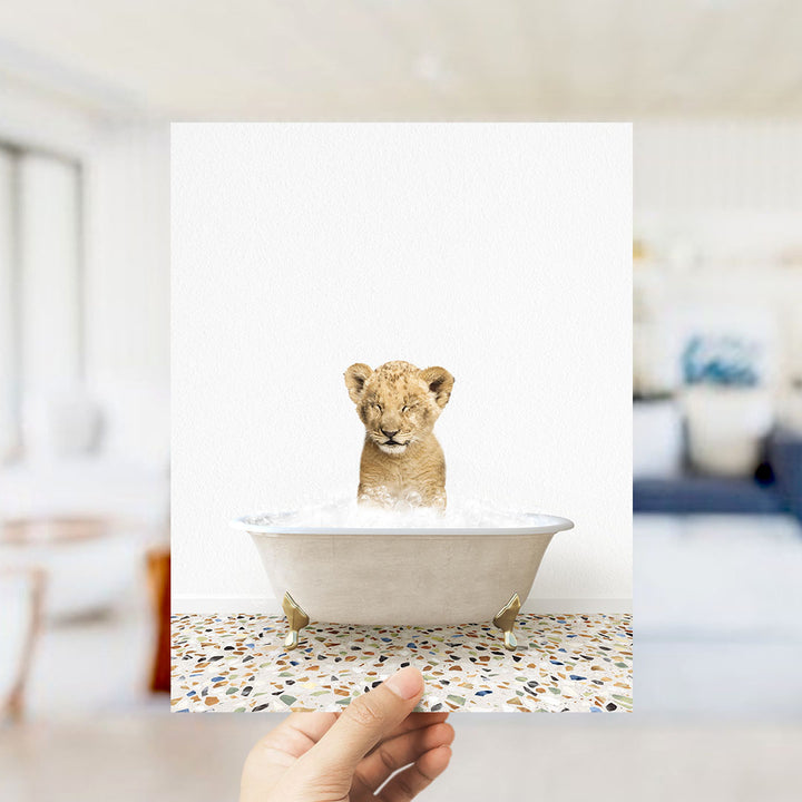 A hand holds up a framed image of a young lion cub sitting in a bathtub filled with water, looking at the camera.