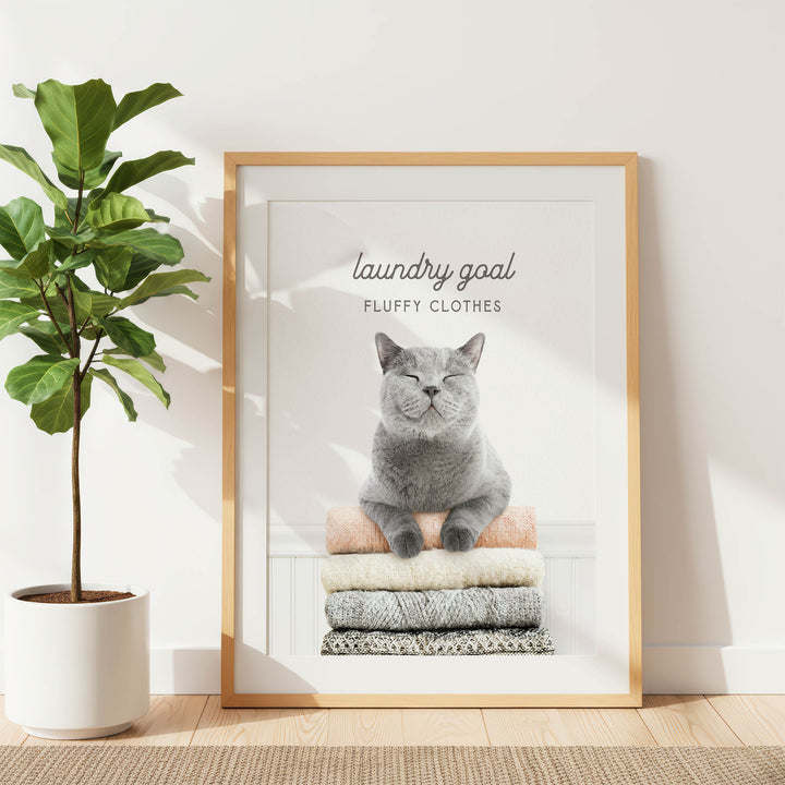 A framed poster with a cat sitting on a stack of clothes, with the text "laundry goal FLUFFY CLOTHES" written above the cat.