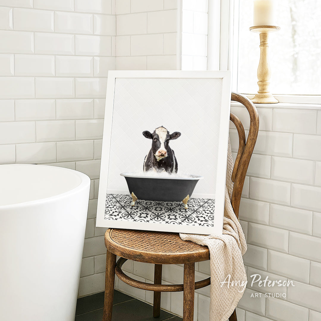 A framed image of a cow sitting in a bathtub, surrounded by a patterned tile floor, is displayed on a wooden chair in a bathroom setting.