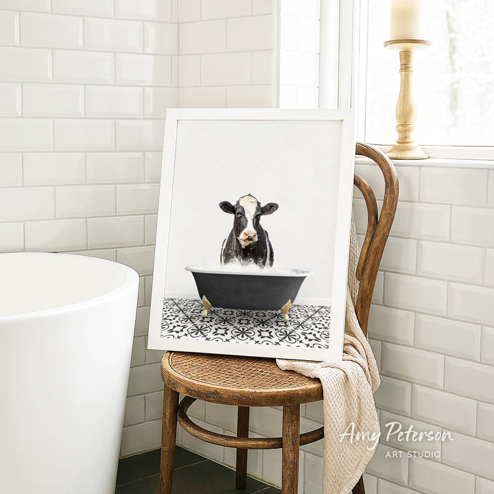 A framed image of a cow sitting in a bathtub, surrounded by a patterned tile floor, is displayed on a wooden chair in a bathroom setting.