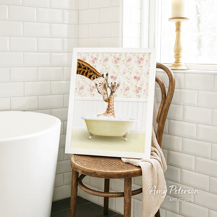 A framed image of two giraffes sitting in a bathtub, with one giraffe reaching over the other, is displayed on a wooden chair in a bathroom setting.
