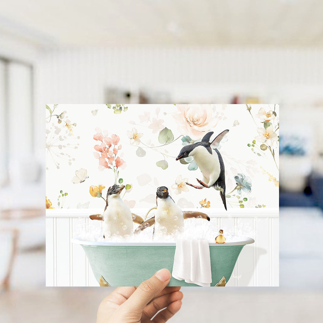 A hand holds up a framed image depicting two penguins sitting in a bathtub filled with bubbles, surrounded by a floral pattern.