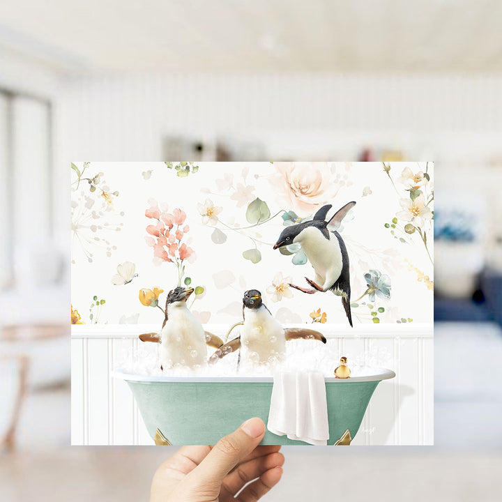 A hand holds up a framed image depicting two penguins sitting in a bathtub filled with bubbles, surrounded by a floral pattern.