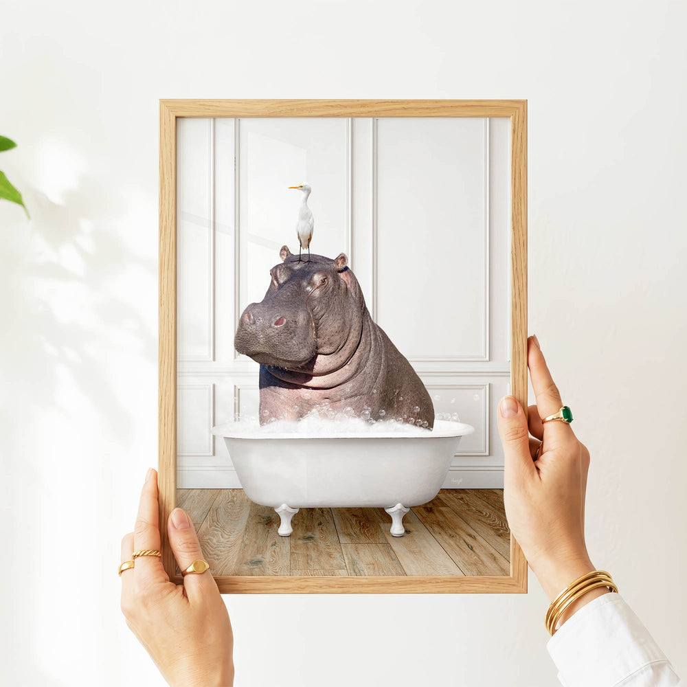 A person's hands are holding a picture frame containing a photograph of a hippopotamus sitting in a bathtub, with a seagull perched on its head.