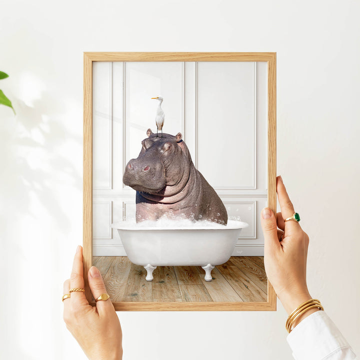 A person's hands are holding a picture frame containing a photograph of a hippopotamus sitting in a bathtub, with a seagull perched on its head.