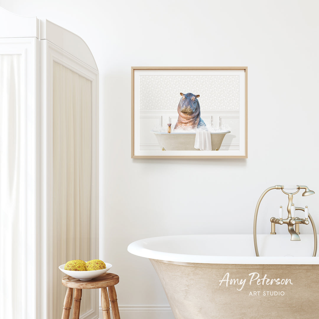 A framed image of a hippopotamus sitting in a bathtub is hanging on a white wall above a freestanding bathtub in a bathroom.