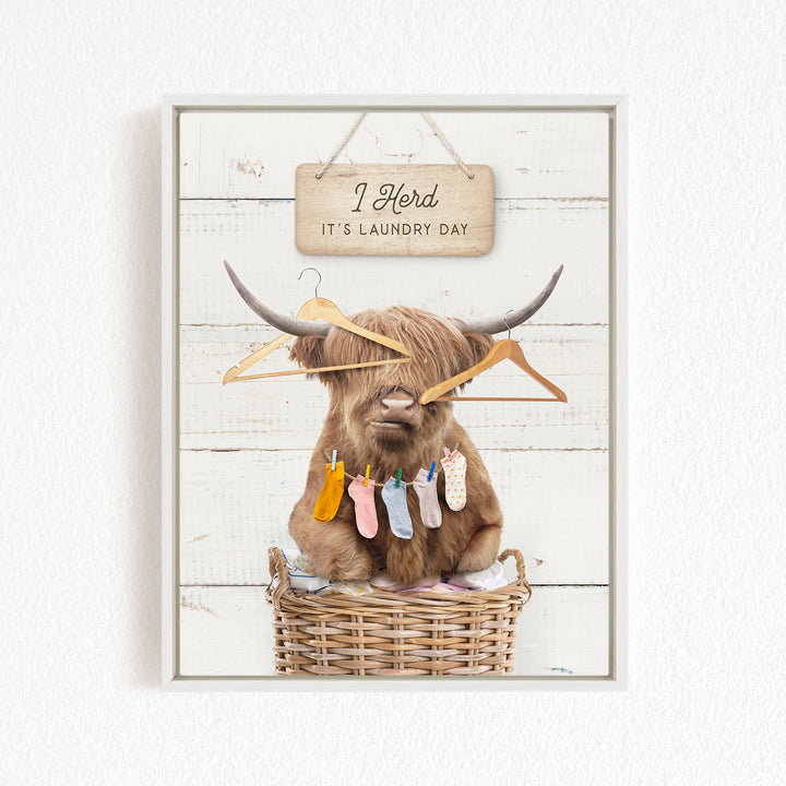 A brown cow with horns is sitting in a wicker basket, surrounded by clothes and hanging clothes on a clothesline. Above the basket, there is a wooden sign that says "I herd it's laundry day".