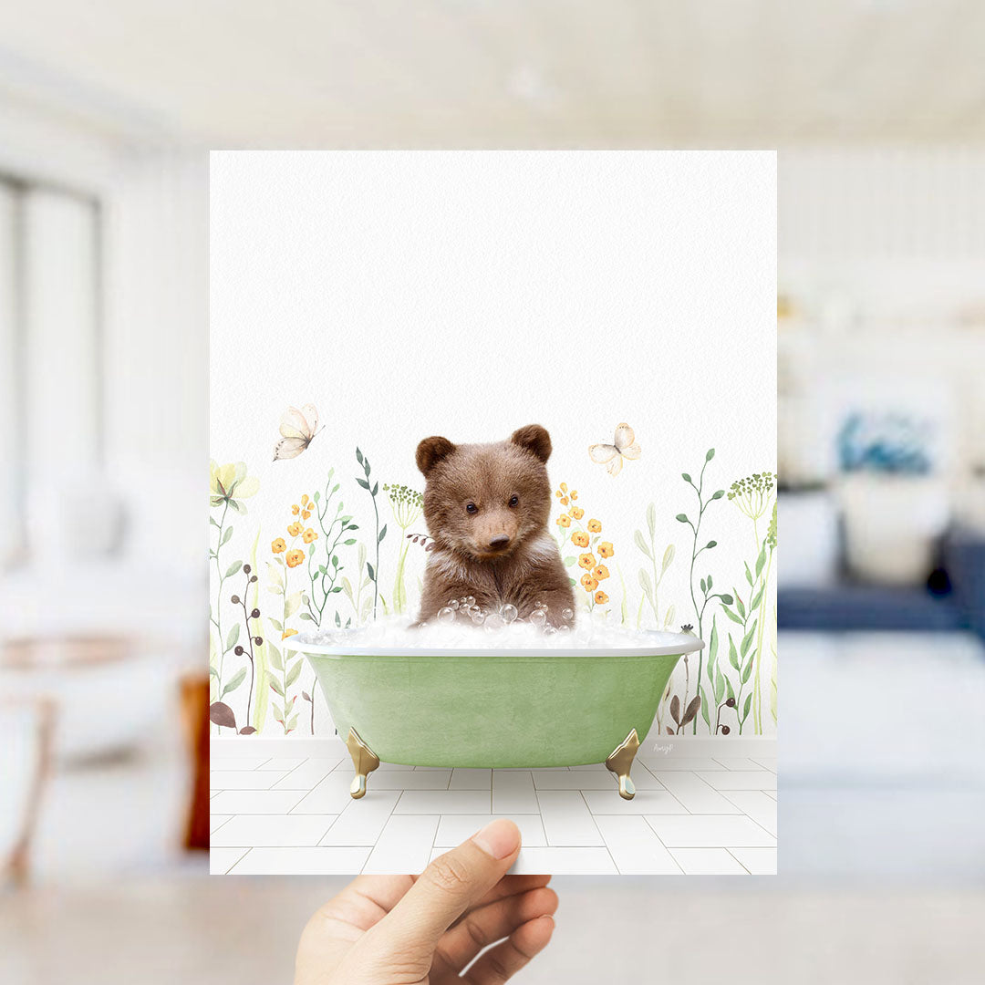 A hand is holding up a framed image of a small brown bear sitting in a green bathtub filled with bubbles, surrounded by flowers and butterflies.