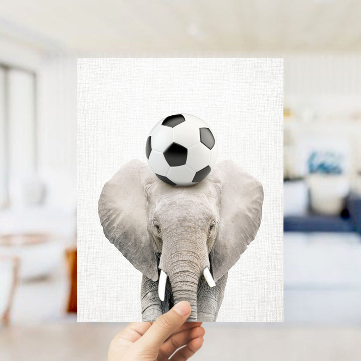 A hand holds up a framed image of an elephant balancing a soccer ball on its head.