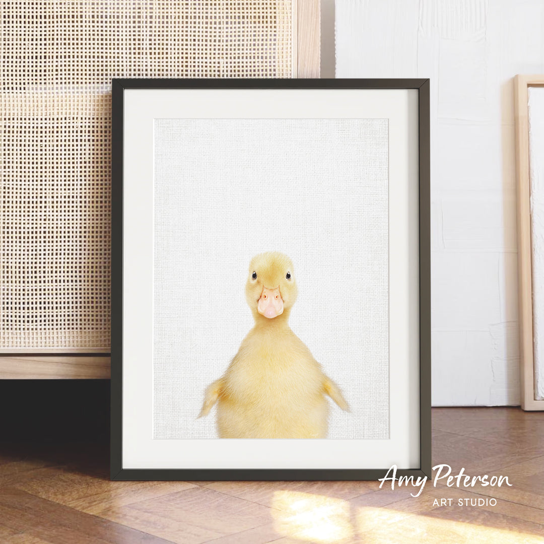 A framed photograph of a cute yellow duck with a playful expression, placed on a wooden floor against a white wall.