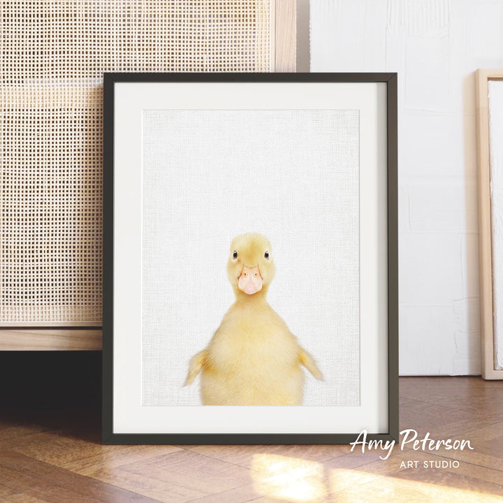 A framed photograph of a cute yellow duck with a playful expression, placed on a wooden floor against a white wall.