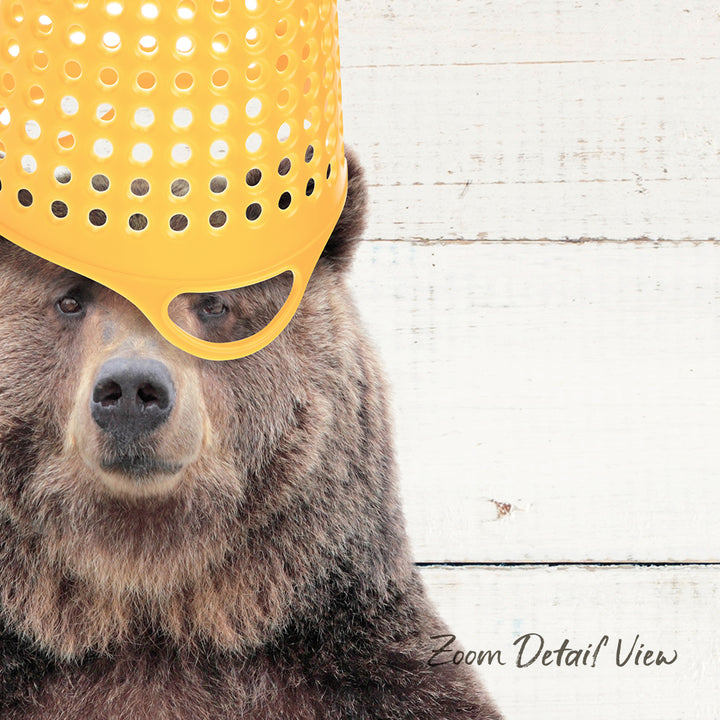 A close-up of a brown bear wearing a yellow plastic bucket hat against a white wooden background.