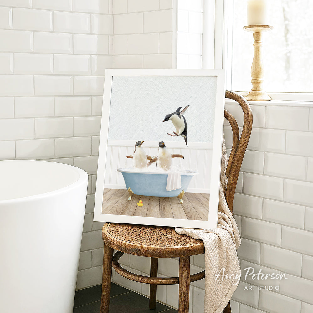 A framed image of two penguins sitting in a bathtub, with one penguin standing on its hind legs, is displayed on a wooden chair in a bathroom setting.