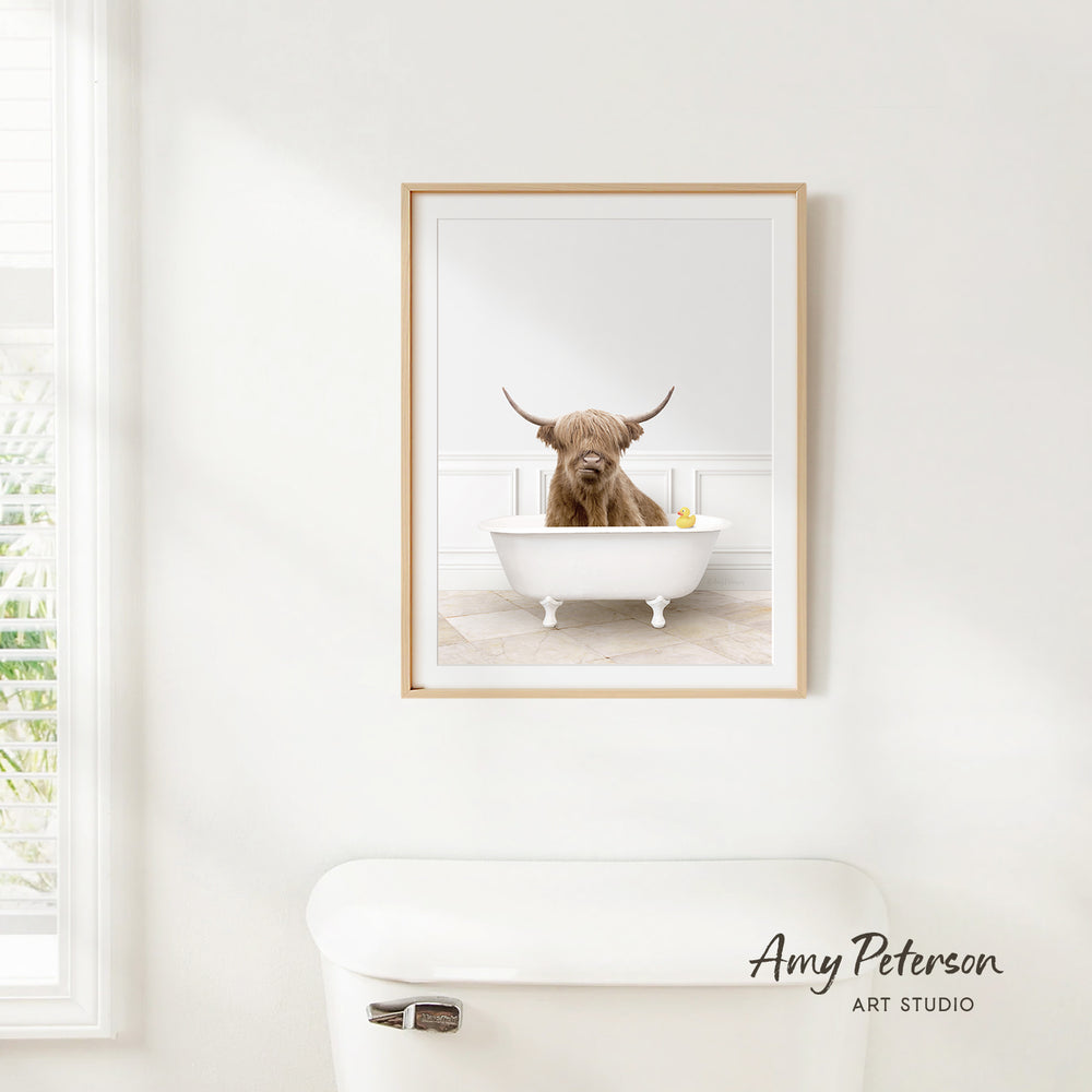 A framed image of a Highland cow sitting in a bathtub, with a window and toilet visible in the background.