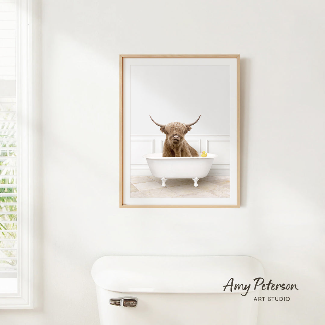 A framed image of a Highland cow sitting in a bathtub, with a window and toilet visible in the background.