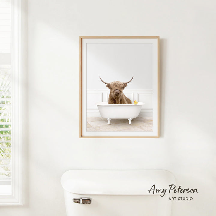 A framed image of a Highland cow sitting in a bathtub, with a window and toilet visible in the background.