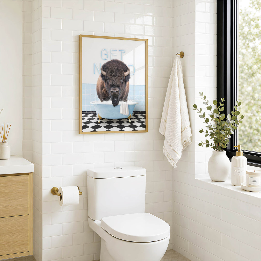 a bathroom with a white toilet, a framed picture of a buffalo in a bathtub, and a towel hanging on a rack.