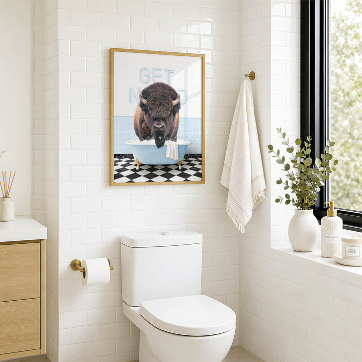 a bathroom with a white toilet, a framed picture of a buffalo in a bathtub, and a towel hanging on a rack.