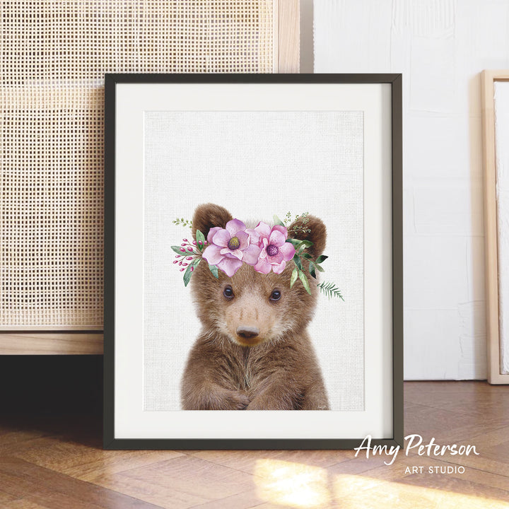 A framed print of a cute bear wearing a floral crown, with a wooden floor and a woven basket in the background.