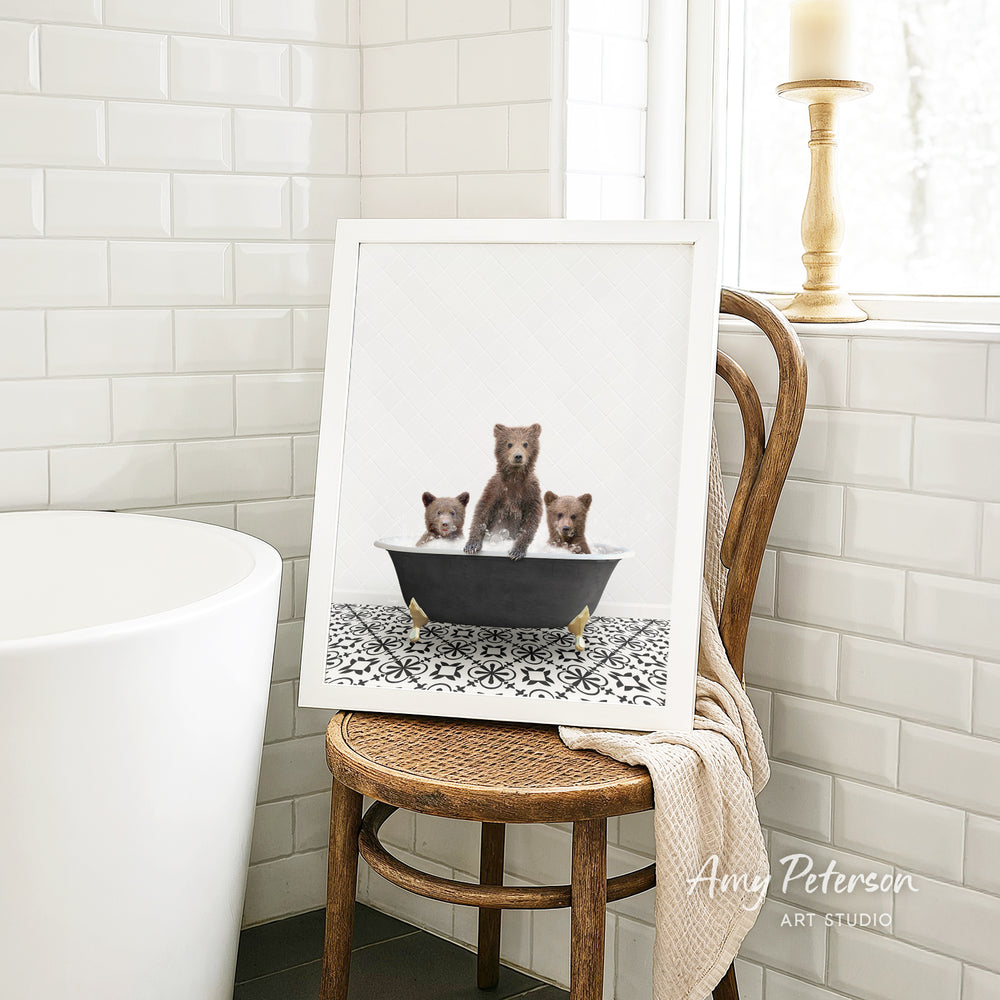 A framed image of three bears sitting in a bathtub, with one bear appearing to be a baby bear. The bears are sitting on a black bathtub with a white patterned tile floor. The image is displayed on a wooden chair in a bathroom setting.