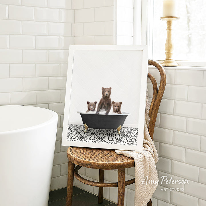 A framed image of three bears sitting in a bathtub, with one bear appearing to be a baby bear. The bears are sitting on a black bathtub with a white patterned tile floor. The image is displayed on a wooden chair in a bathroom setting.