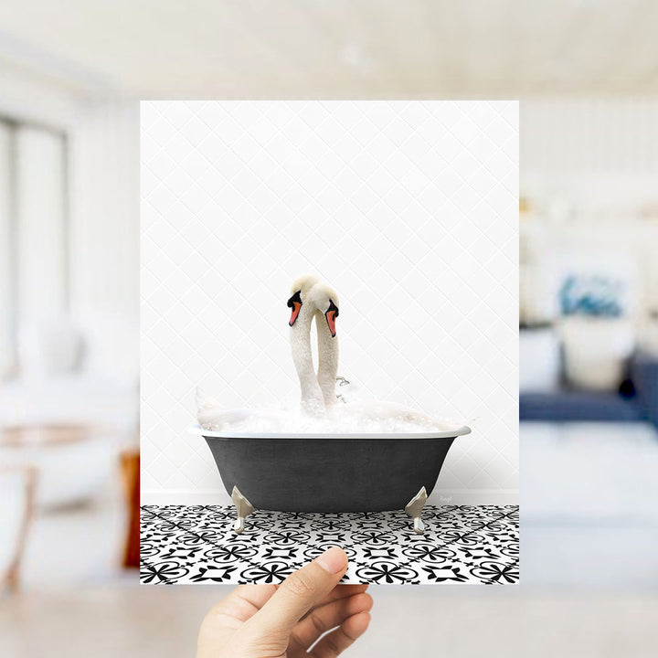 A hand holds up a framed photograph of two swans standing in a bathtub filled with water, creating a whimsical and playful scene.